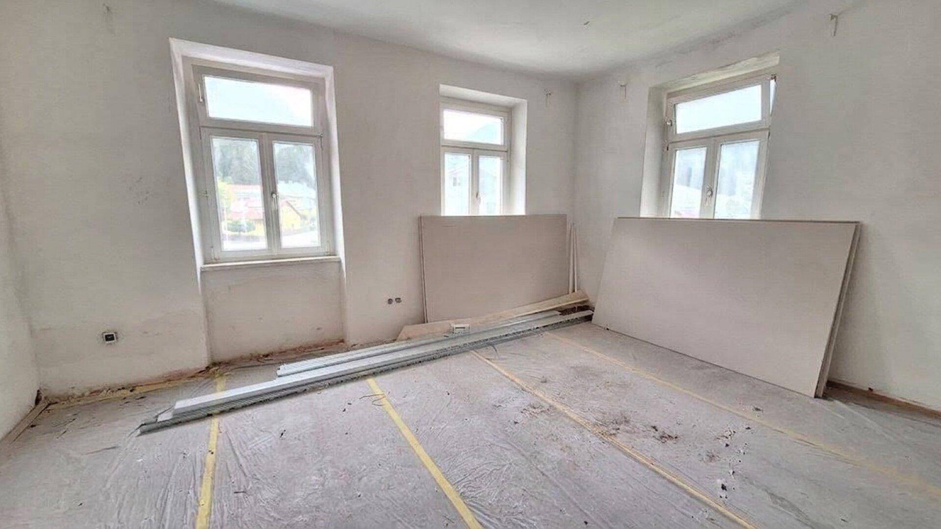 Renovierungsbedürftiger Raum mit drei Fenstern und vorbereitetem Boden für neue Beläge.