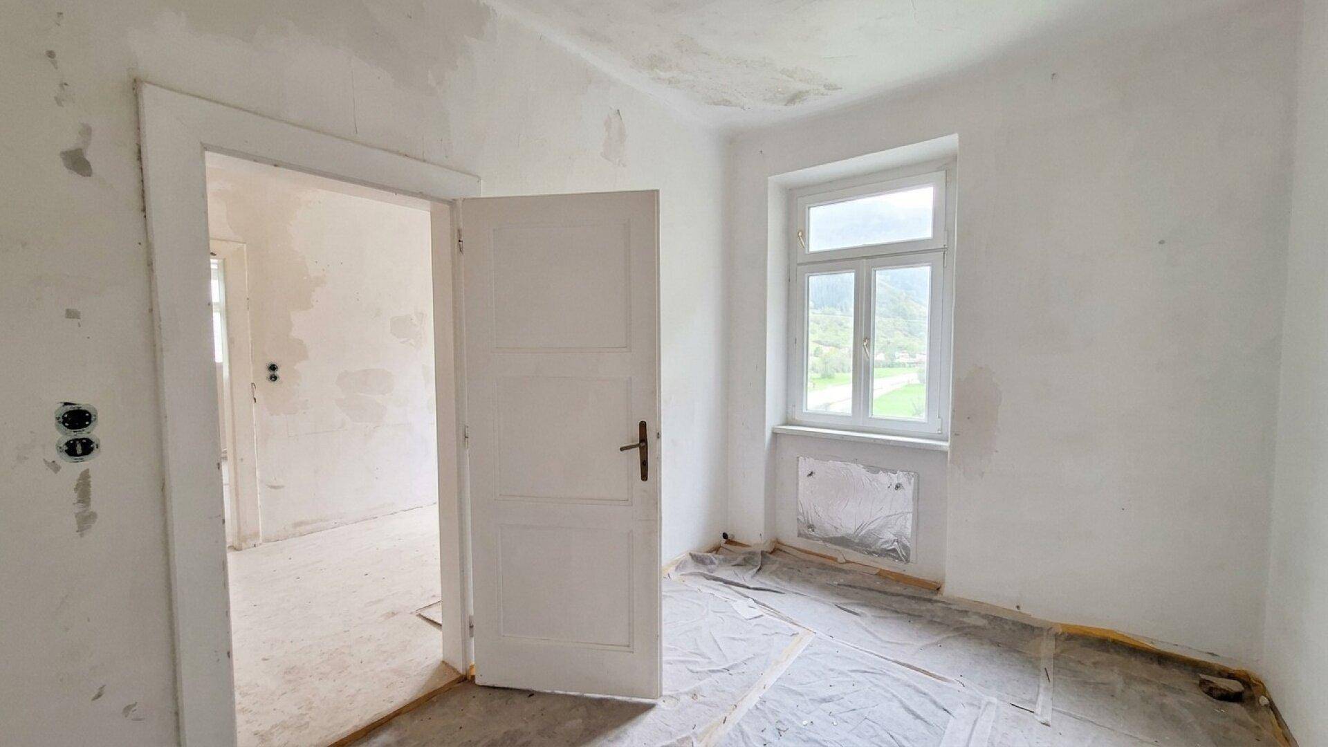Leerer Raum in Renovierung mit einem Fenster und offener Tür zu einem weiteren Bereich.