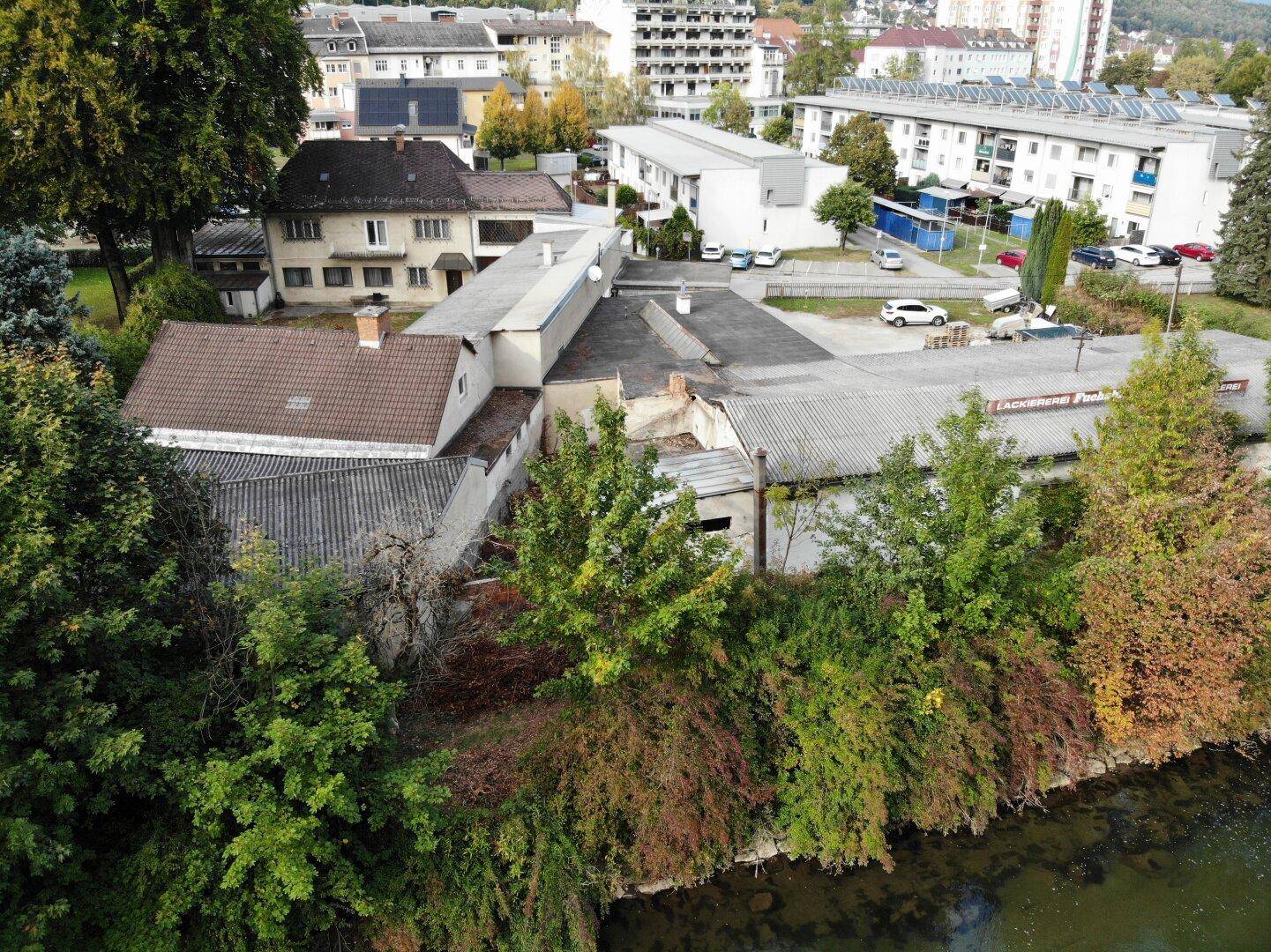Gebäudekomplex mit verschiedenen Dachtypen, umgeben von Bäumen und einem Fluss im Vordergrund.
