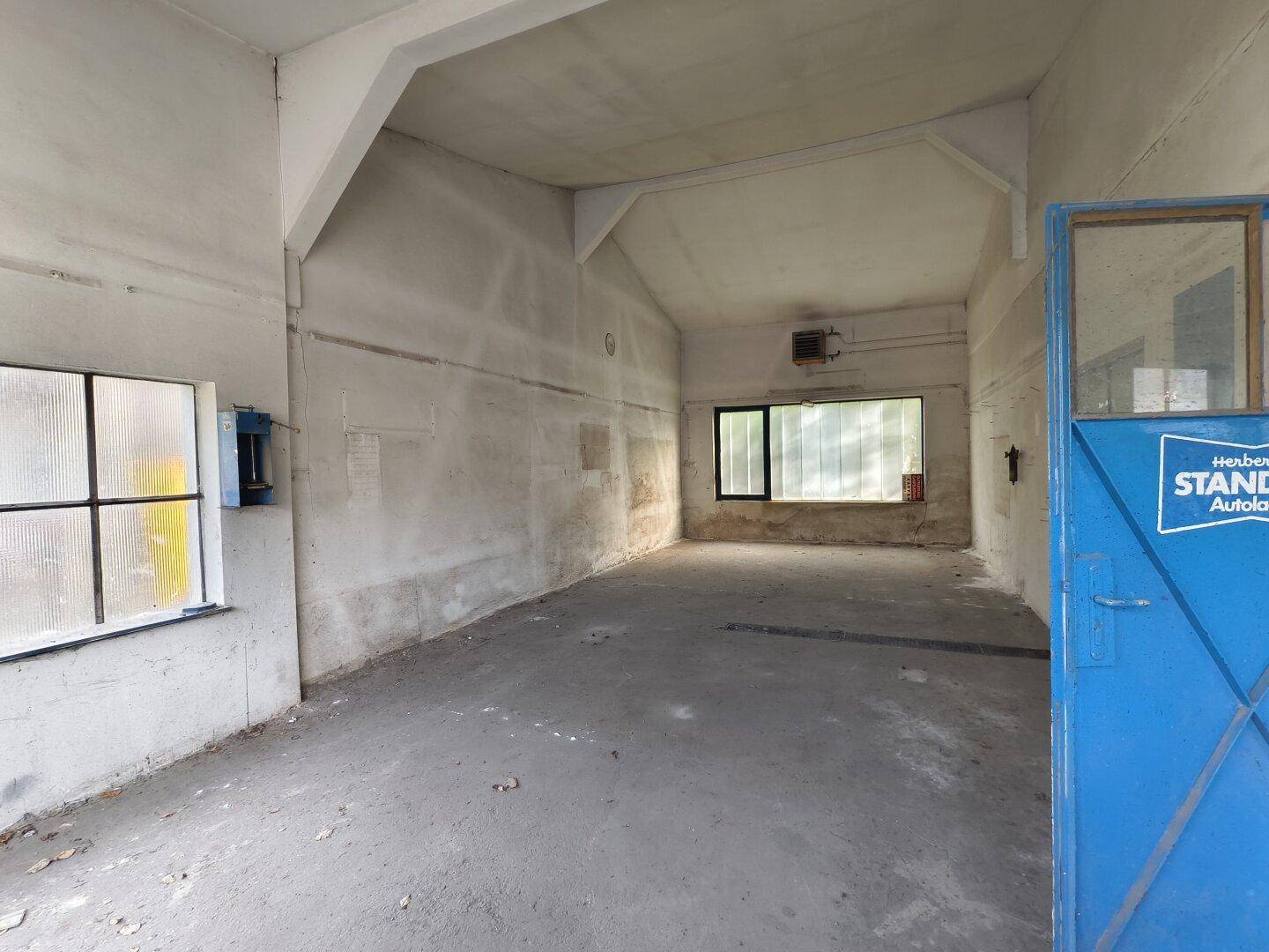 Kleiner, leerer Raum mit Betonboden, weißen Wänden und einem Fenster, mit blauer Tür.