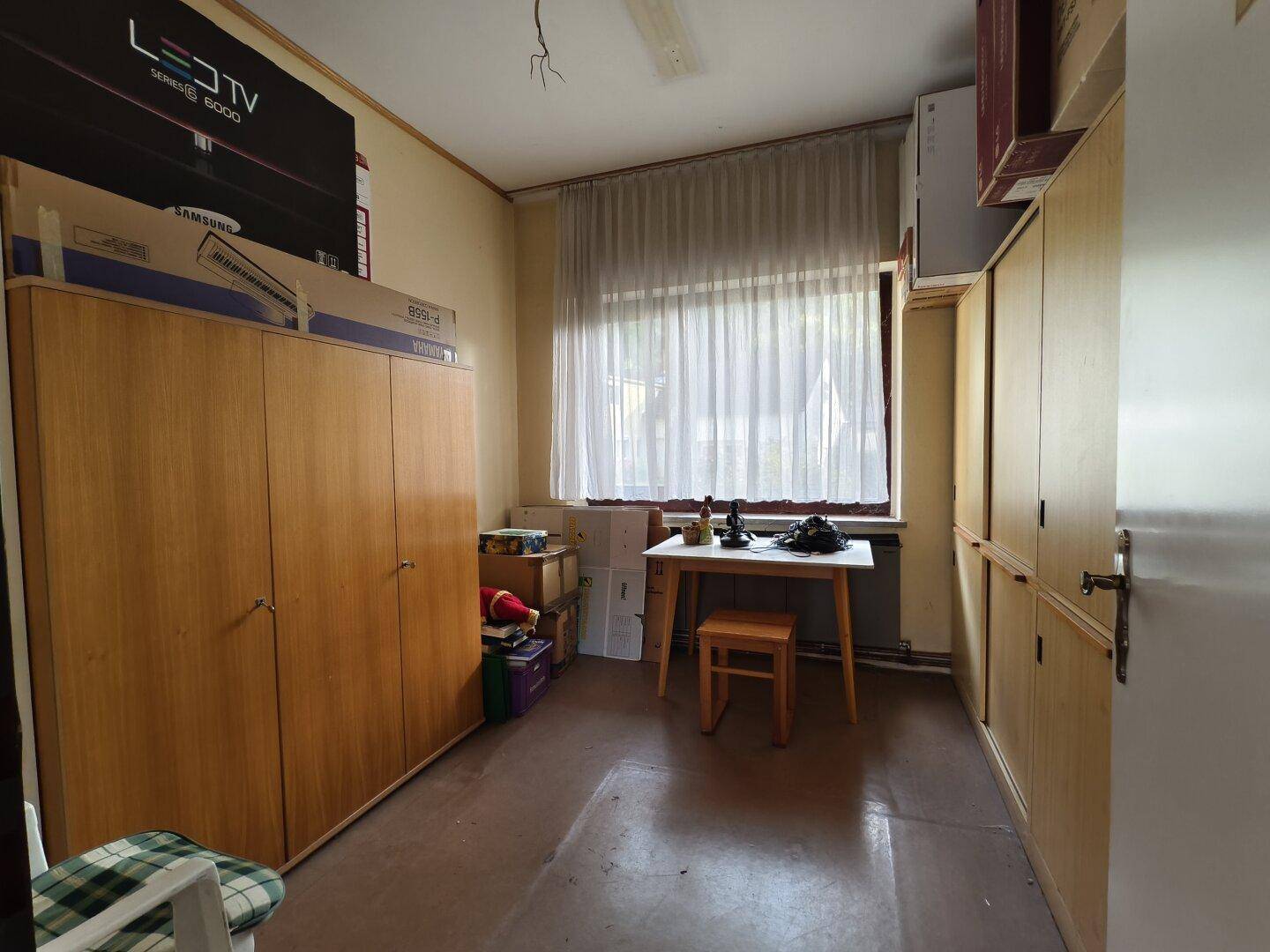 Kleines Zimmer mit Fenster, Vorhängen und eingebauten Holzschränken, möbliert mit Tisch und Stuhl.
