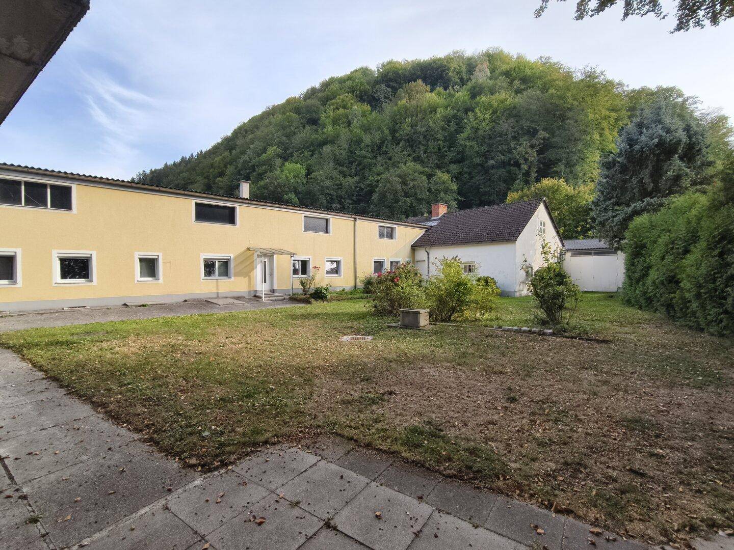 Gelbes und weißes Gebäude mit Rasenfläche und bewaldetem Hügel im Hintergrund.