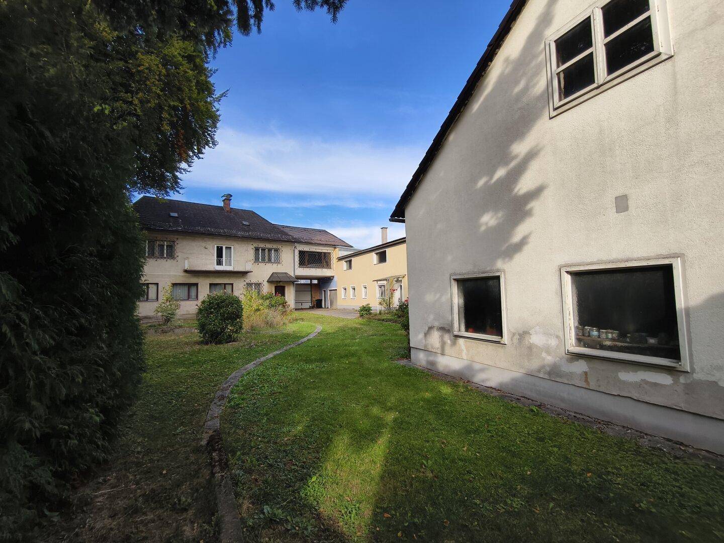 Zwei Gebäude, eines hell, das andere gelb, getrennt durch einen grünen Rasenweg unter blauem Himmel.