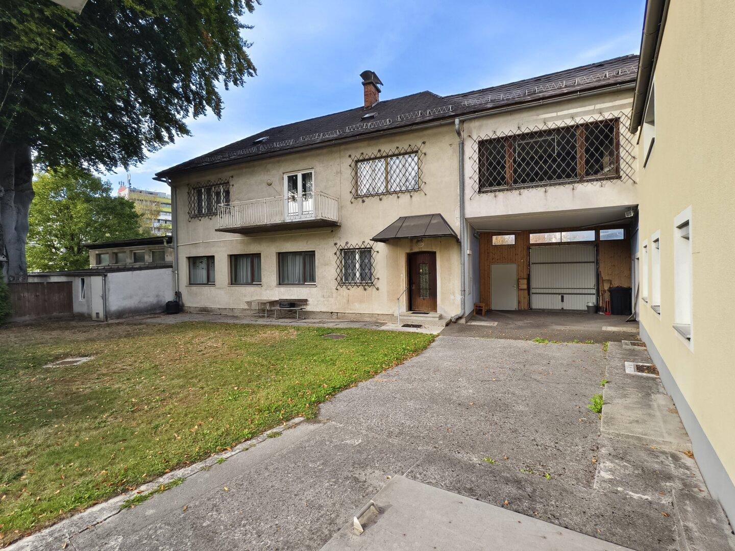 Helles Gebäude mit kleinem Balkon, Garageneinfahrt und gepflastertem Bereich mit Rasenfläche.