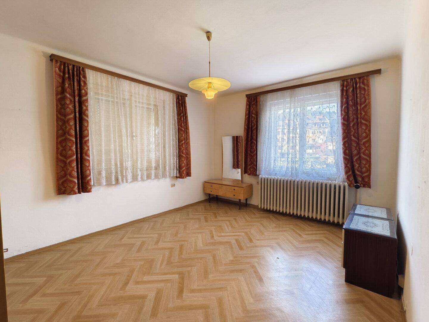 Geräumiges Wohnzimmer mit Fischgrätparkett, zwei Fenstern und traditionellen Vorhängen.