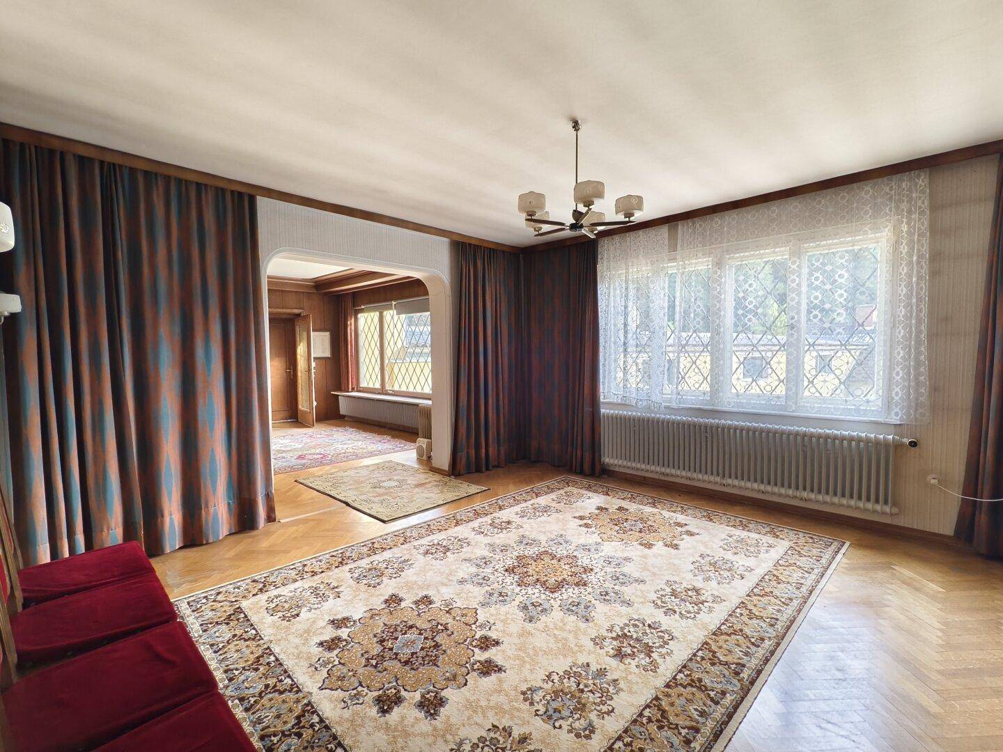 Helles Wohnzimmer mit Parkettboden, großem Teppich und Fenstern mit Spitzenvorhängen.