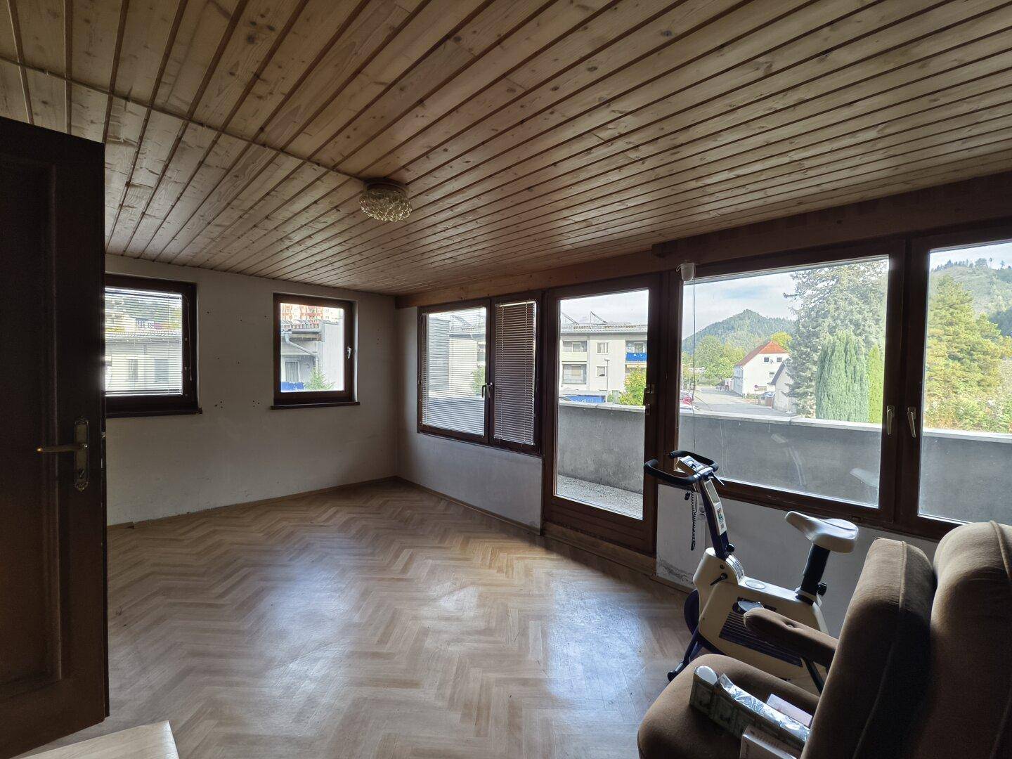 Heller Raum mit Holzdecke, Parkettboden und großen Fenstern mit Ausblick.
