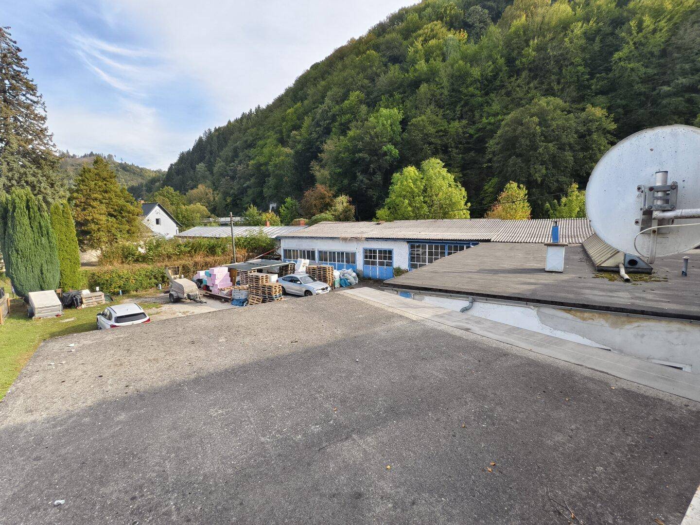 Außenbereich mit Parkplätzen, Gebäuden und bewaldetem Hügel im Hintergrund.