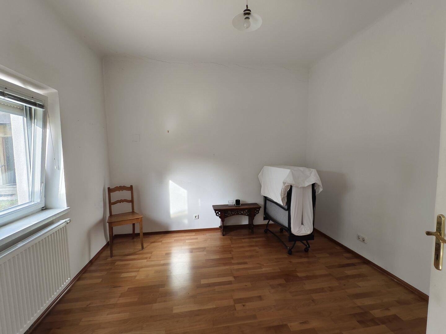 Helles Schlafzimmer mit Holzfußboden, Fenster, Heizkörper und Babybett.