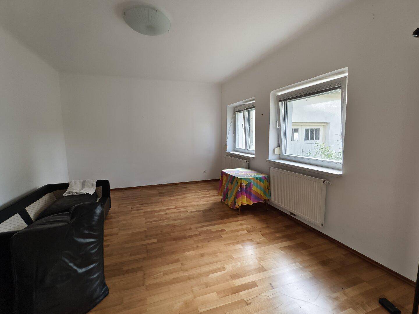 Helles Wohnzimmer mit Holzfußboden, zwei Fenstern und schwarzem Sofa.