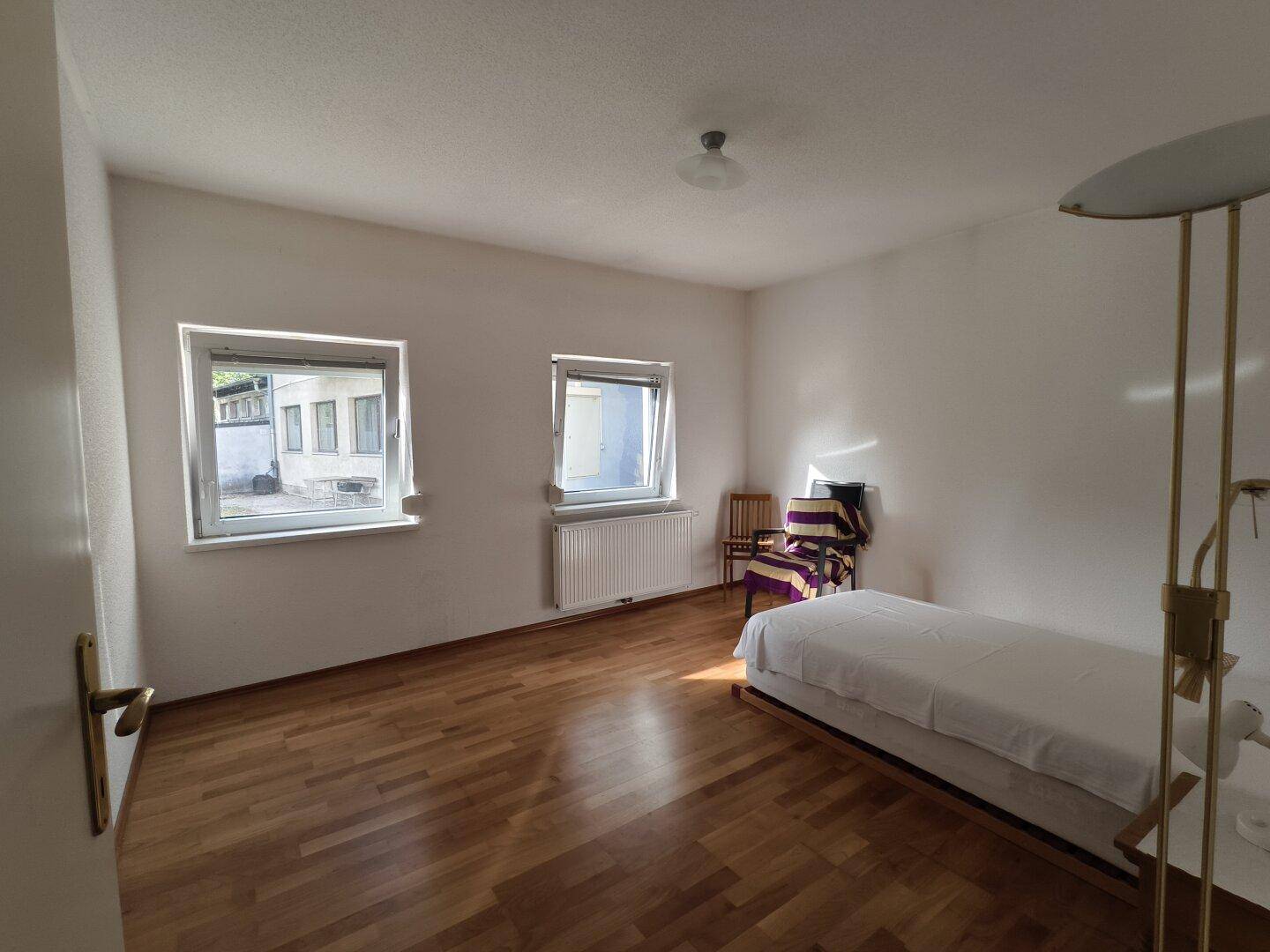Helles Schlafzimmer mit Holzfußboden, weißen Wänden und zwei Fenstern.