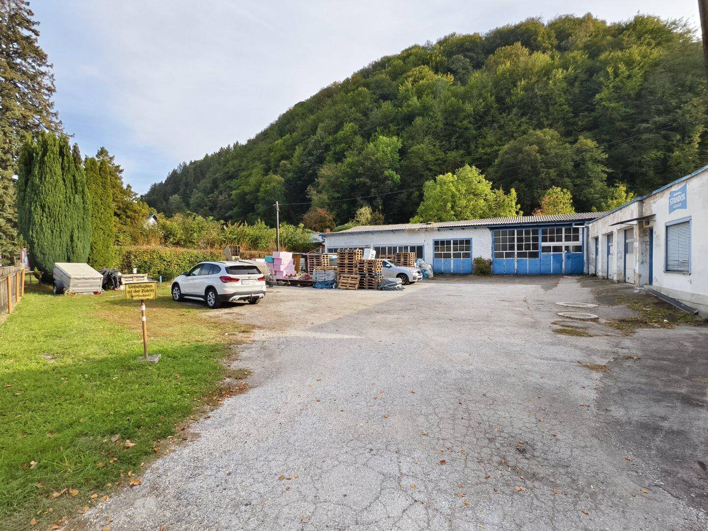 Außenbereich mit großem Parkplatz, mehreren Gebäuden und bewaldetem Hügel im Hintergrund.
