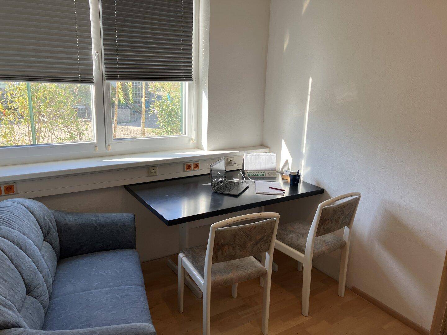Kleiner Bürobereich mit Wandtisch, zwei Stühlen, einem Sofa und Fenstern mit Jalousien.
