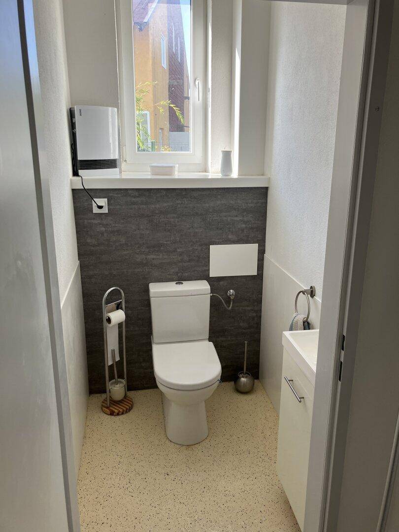 Modernes WC mit wandhängender Toilette, kleinem Waschbecken und Fenster, akzentuiert durch eine graue Wand.