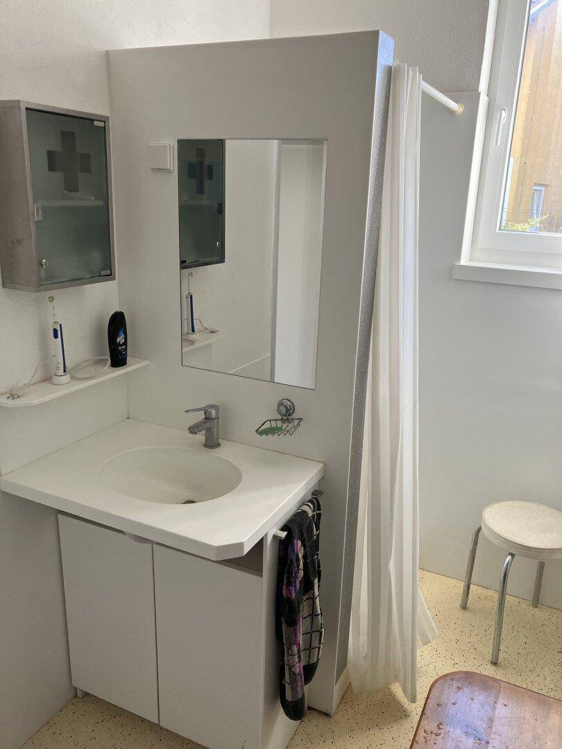 Badezimmer mit Waschtisch, Spiegel und Duschbereich mit Vorhang, hell durch ein Fenster.