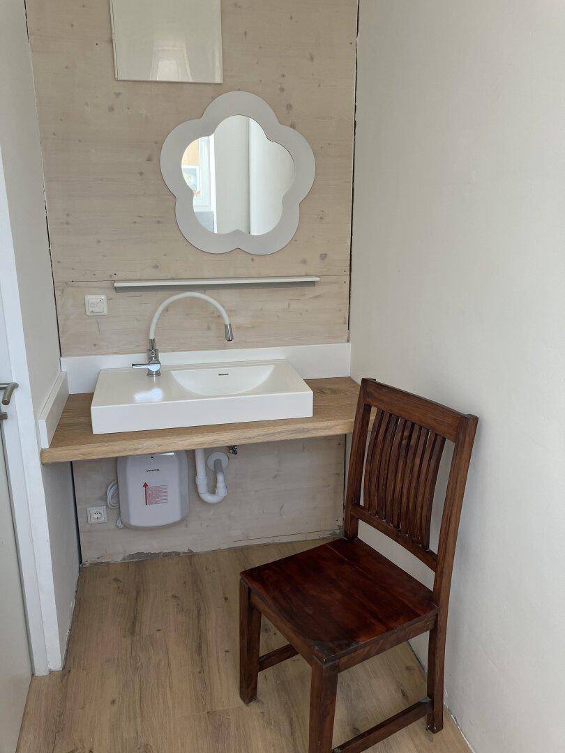 Kleiner Raum mit modernem Waschbecken auf Holzplatte, dekorativem Spiegel und Holzstuhl.