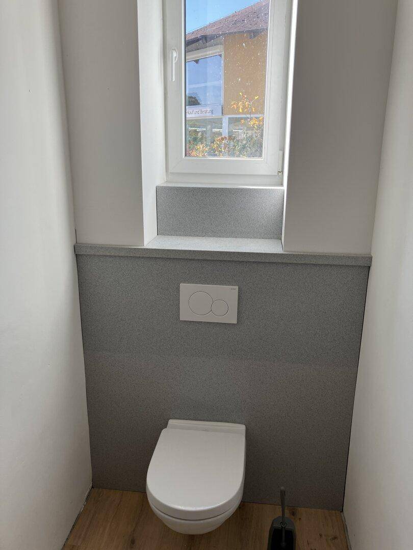 WC-Raum mit wandhängender Toilette und Fenster, betont durch eine graue Akzentwand.