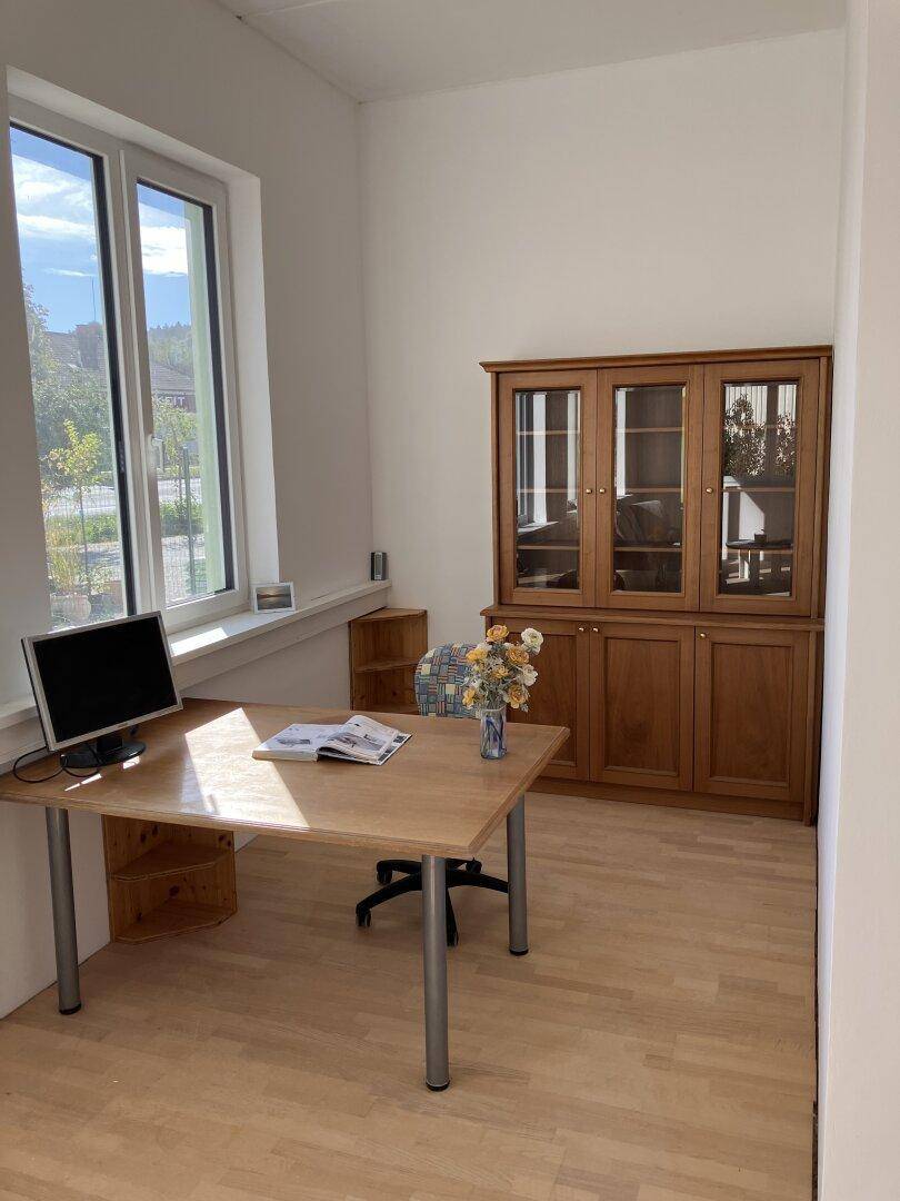 Büroraum mit Holzschreibtisch, Computer und großem Holzvitrinenschrank, hell durch große Fenster.