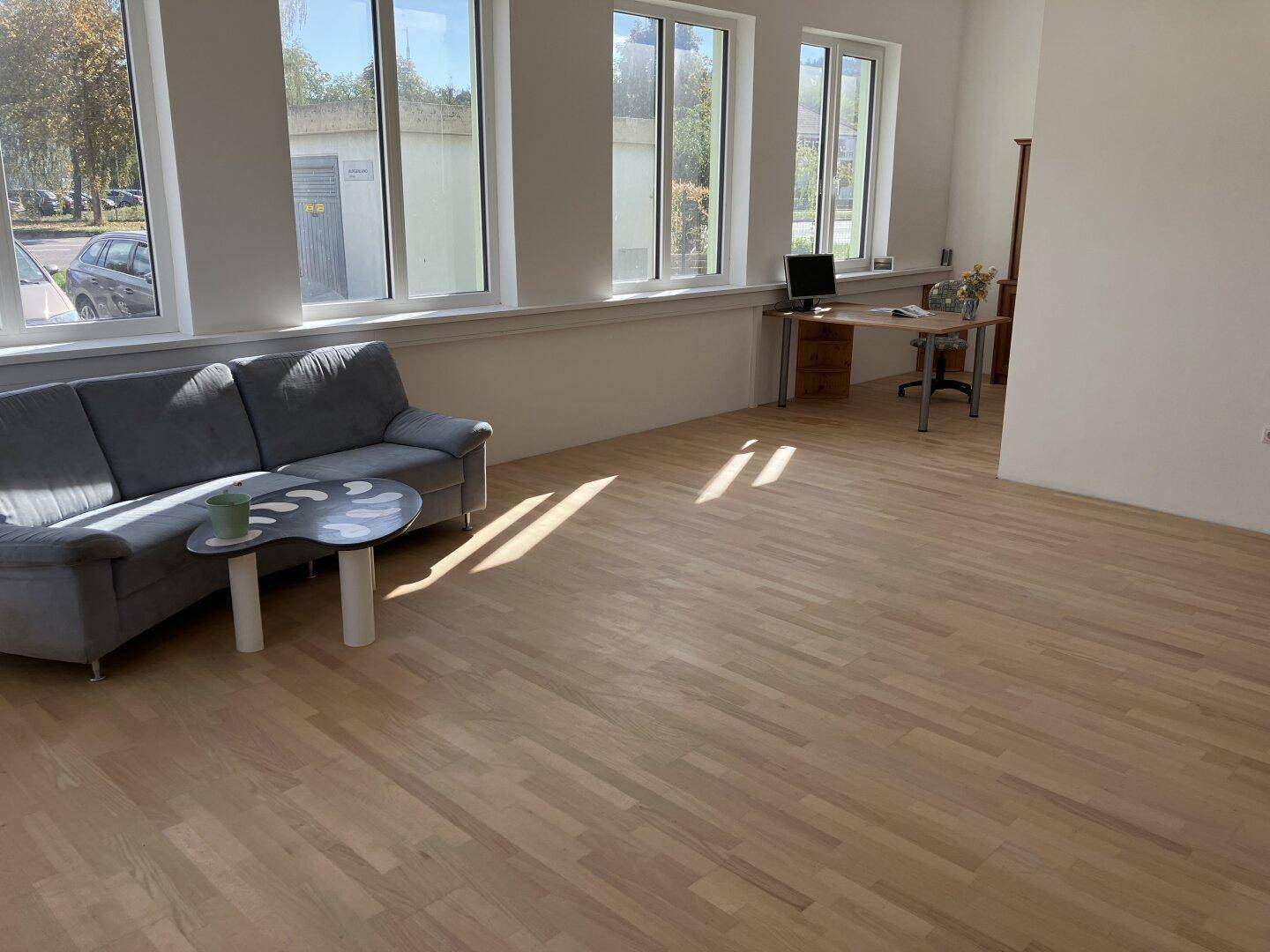 Großer offener Bürobereich mit Sofa, Couchtisch und Schreibtisch, ausgestattet mit Holzboden und großen Fenstern.