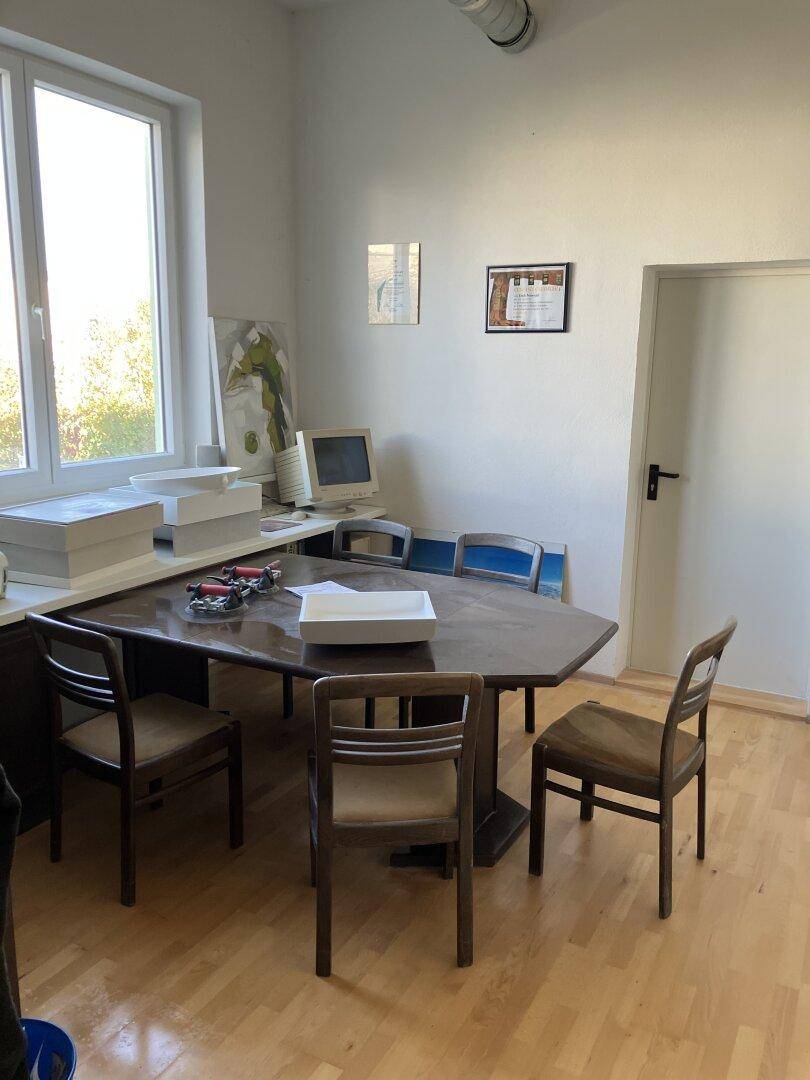 Büroraum mit großem Holztisch, Stühlen und Computerarbeitsplatz, hell durch ein Fenster.