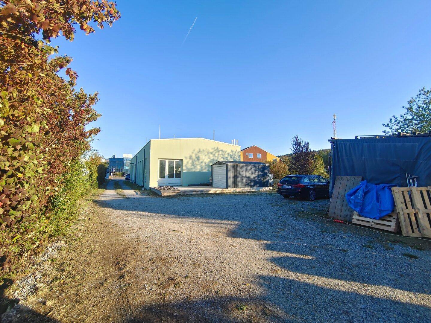 Blick über den Kiesparkplatz auf das Gebäude, einen Schuppen und die umgebende Natur.