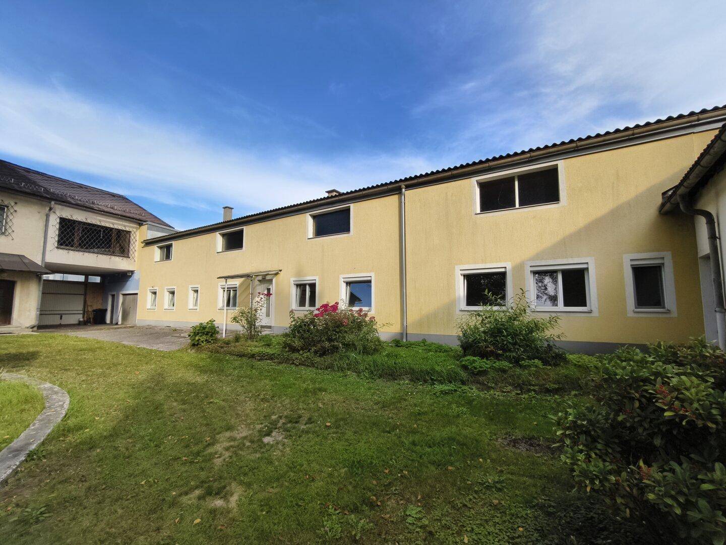 Langes, gelbes Gebäude mit vielen Fenstern und einem kleinen Gartenbereich unter blauem Himmel.