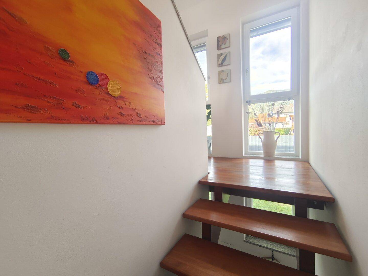 Heller Treppenabsatz mit Holzstufen, weiß gestrichenen Wänden und Fenster mit Ausblick ins Grüne.