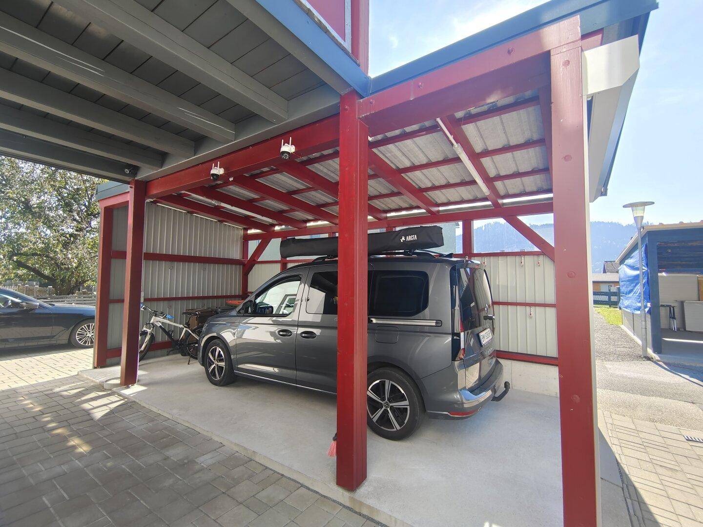 Überdachter Carport mit rotem Holzrahmen und einem geparkten Fahrzeug.