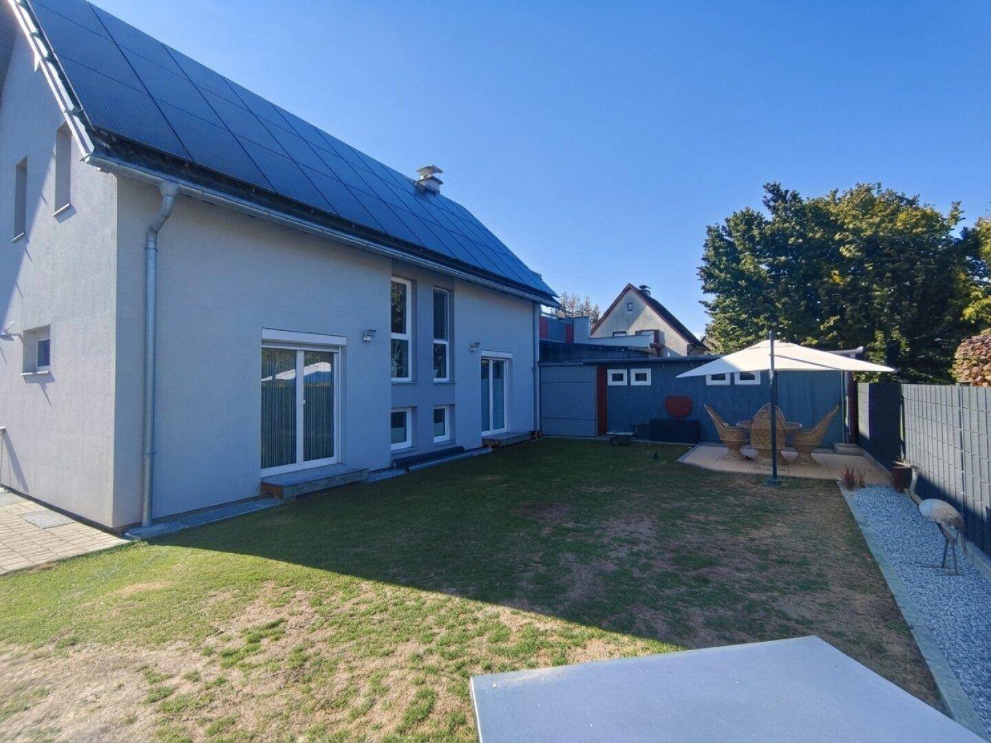 Modernes Haus mit Photovoltaikanlage auf dem Dach und einem gepflegten Garten mit Sonnenschirm.