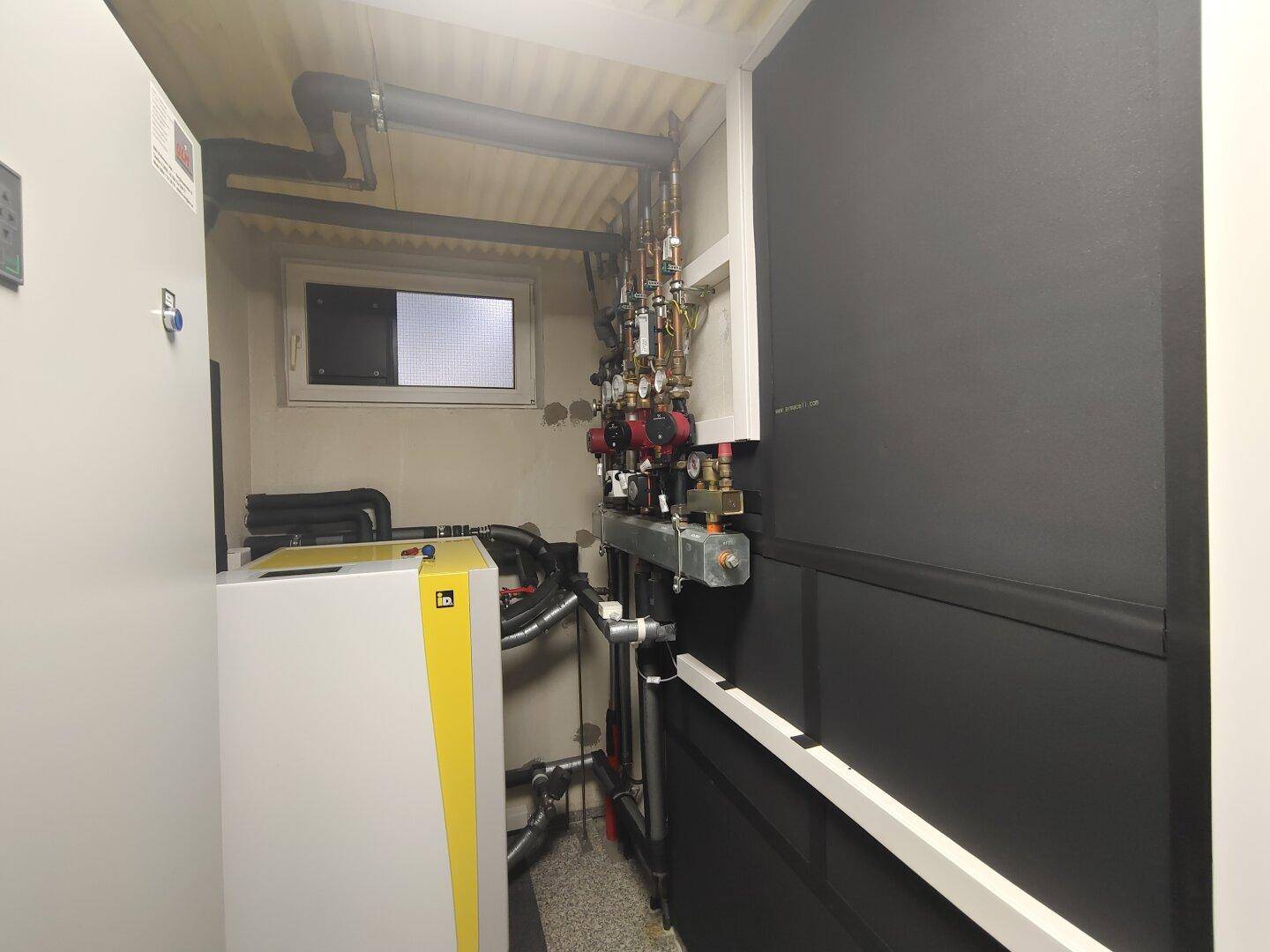 Technikraum im Keller mit Heizungsanlage, Rohren und einem kleinen Fenster.