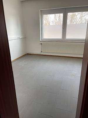 Heller, leerer Raum mit grauen Fliesen, großen Fenstern und Heizkörpern, ideal für Büro oder Schlafzimmer.