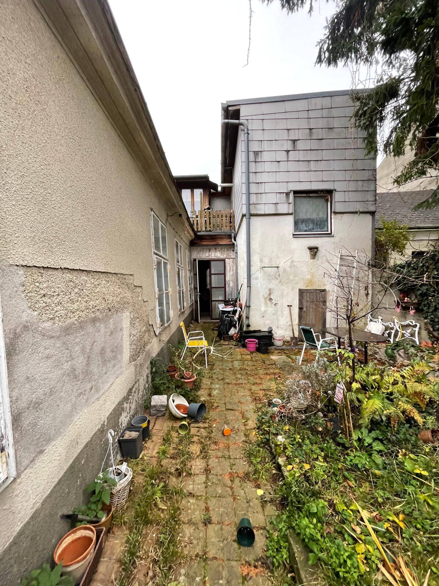 Hinterhofgarten mit gepflastertem Pfad, verschiedenen Pflanzen und Gartenmöbeln, umgeben von Gebäuden.