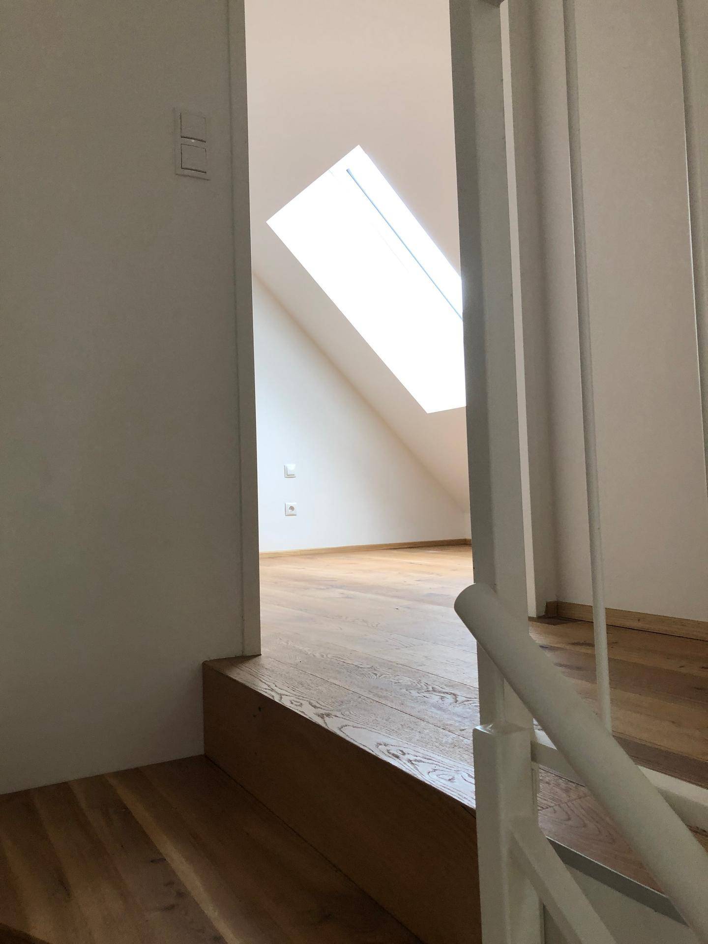 Treppenaufgang mit Blick in ein helles Zimmer im Dachgeschoss mit Holzboden und Dachfenster.