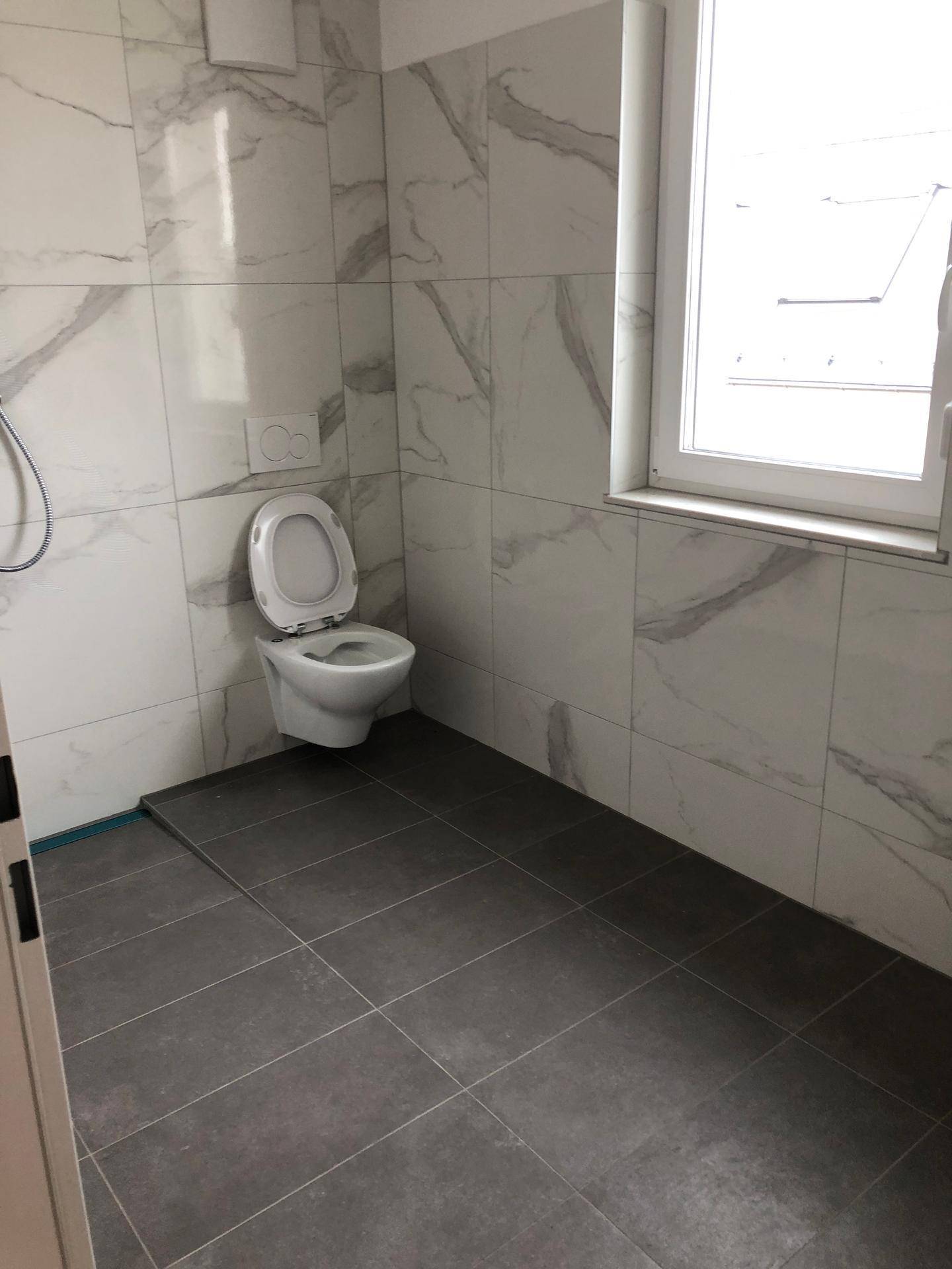 Helles Badezimmer mit wandhängendem WC, Duschbereich und Fenster für natürliche Belüftung.