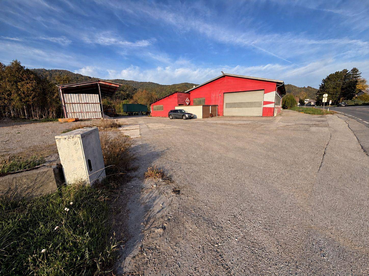 Weitläufige Außenansicht einer roten Industriehalle mit einem geparkten Auto und einer bergigen Landschaft im Hintergrund.