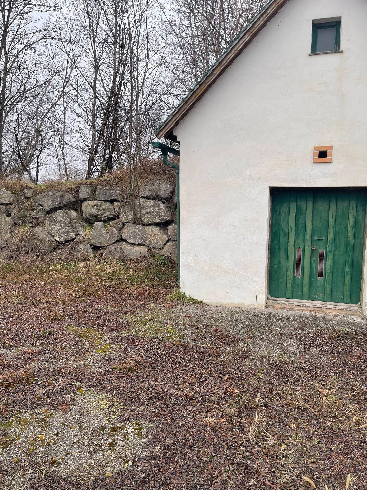 Ländliches Gebäude mit weißer Fassade und grüner Holztür, umgeben von Bäumen und einer Steinmauer.
