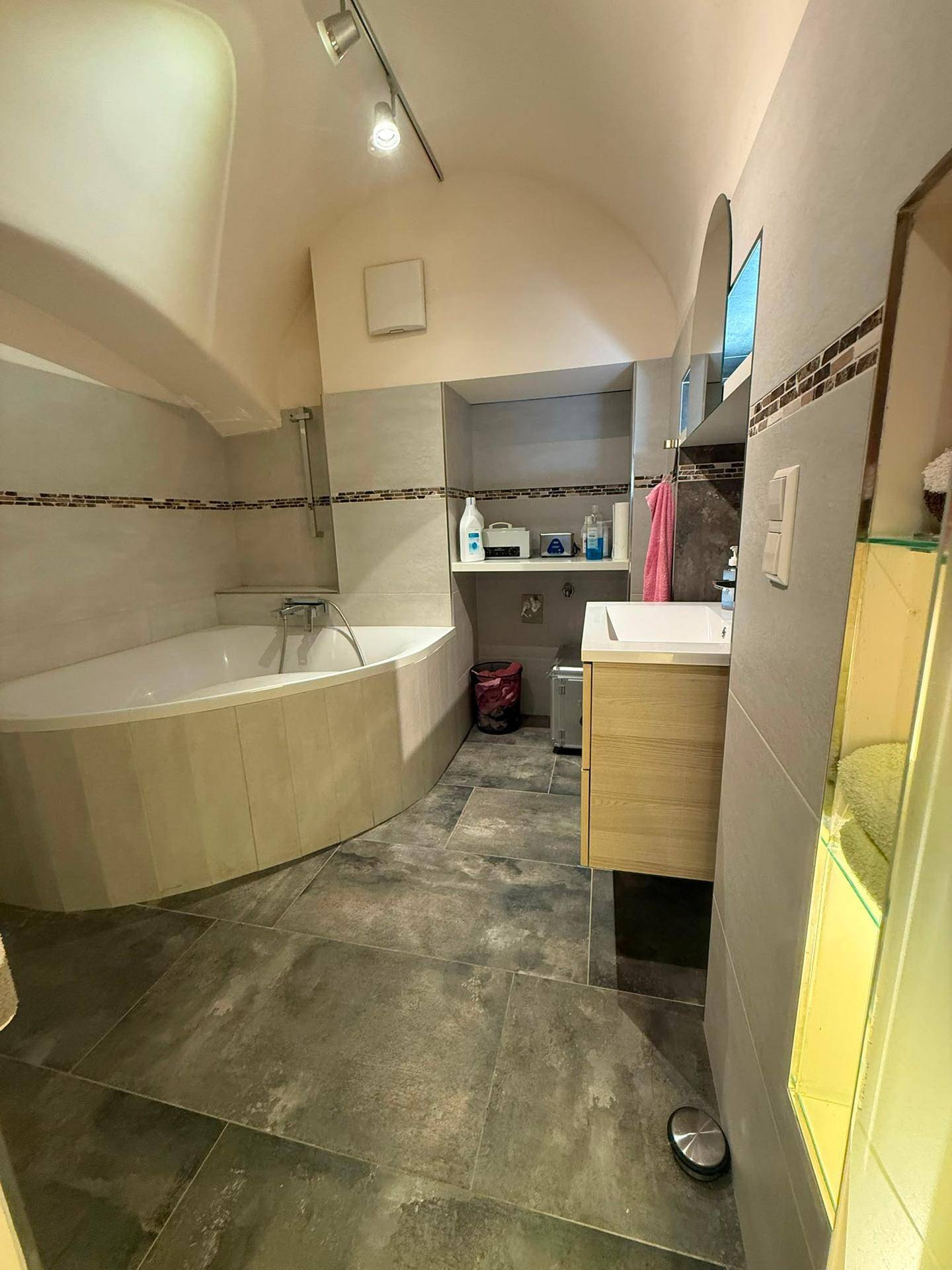 Geräumiges Badezimmer mit Eckbadewanne, modernem Waschtisch und dunklen Fliesen unter gewölbter Decke.