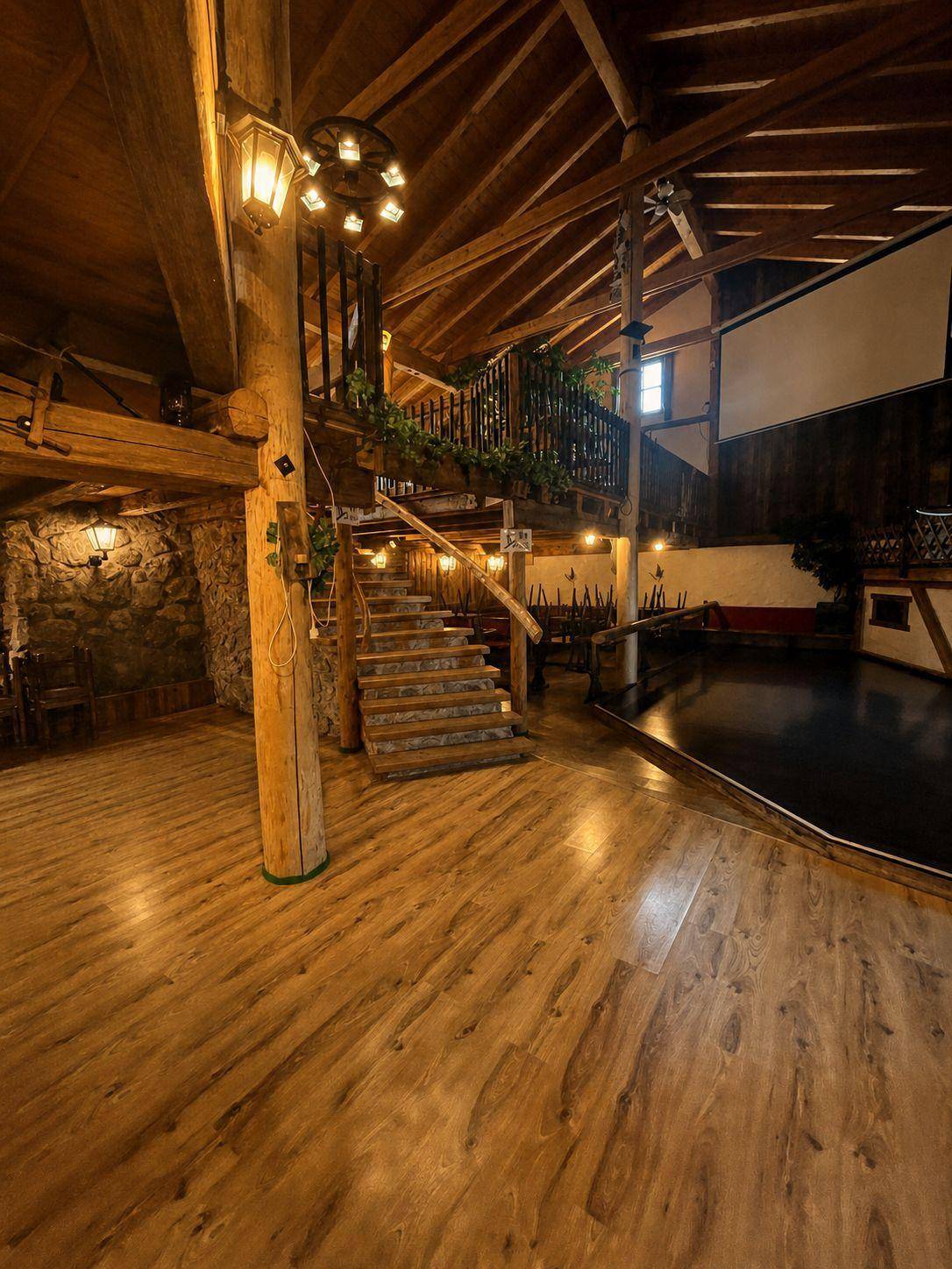 Großer Saal mit Holzboden, Holzbalken und einer geschwungenen Treppe, die zu einer oberen Ebene führt.