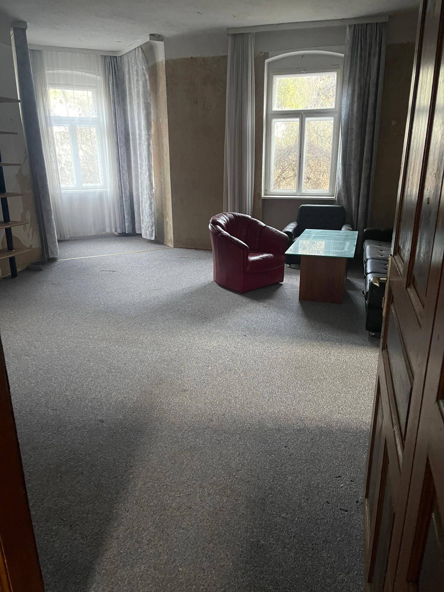 Geräumiges Wohnzimmer mit Teppichboden, zwei Fenstern und einfacher Möblierung in einem älteren Zustand.