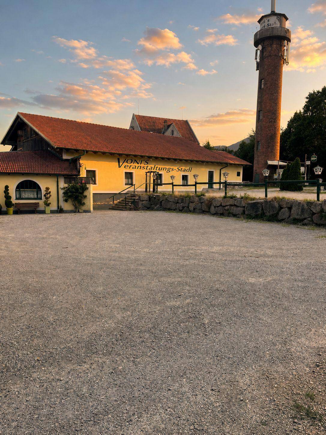 Gebäudekomplex mit Schotterparkplatz und Turm bei Sonnenuntergang, warme Lichtstimmung.