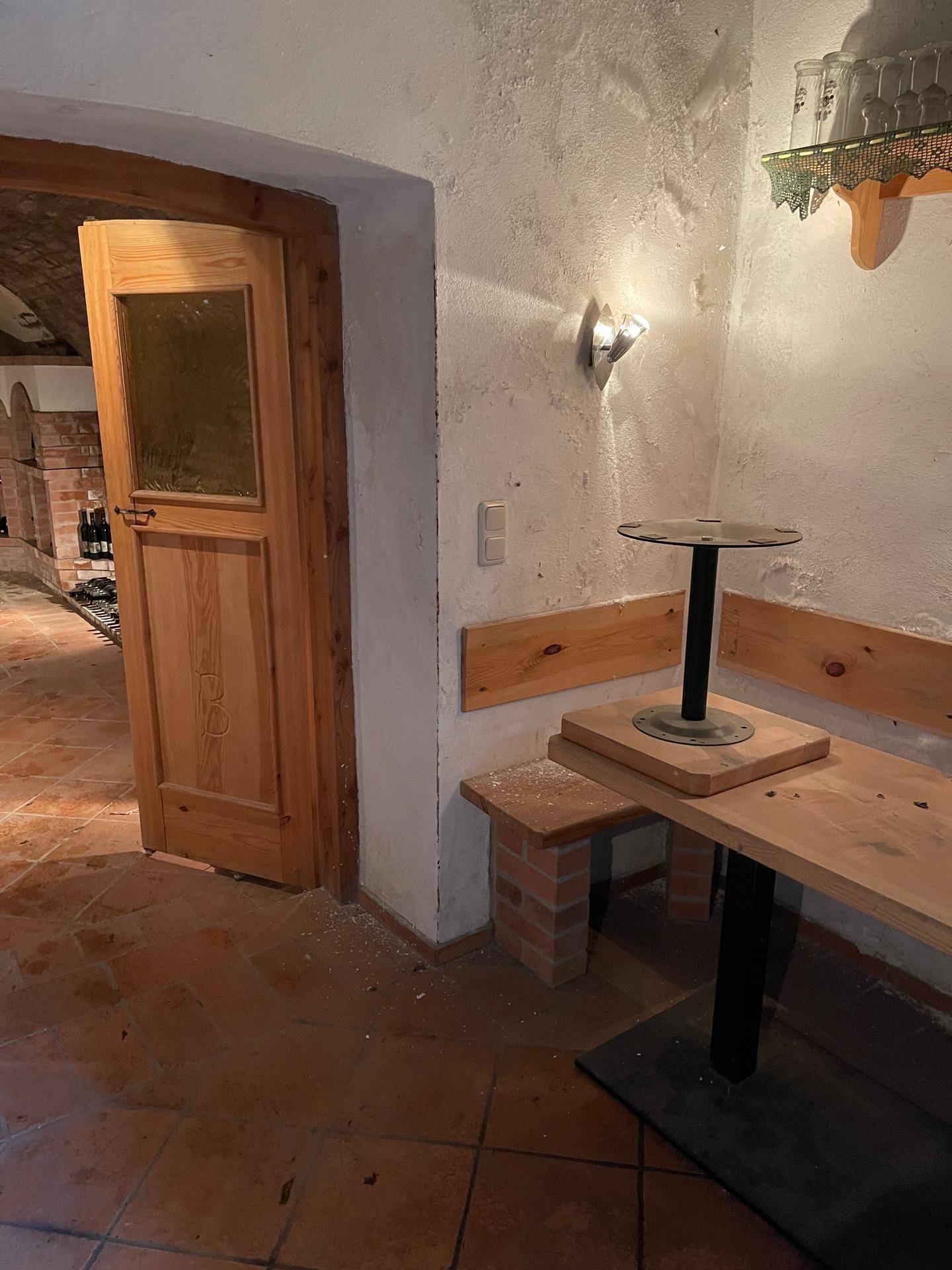 Gemütlicher Sitzbereich in einem rustikalen Kellerraum mit Holzbänken und Terrakottaboden, neben einer Holztür.