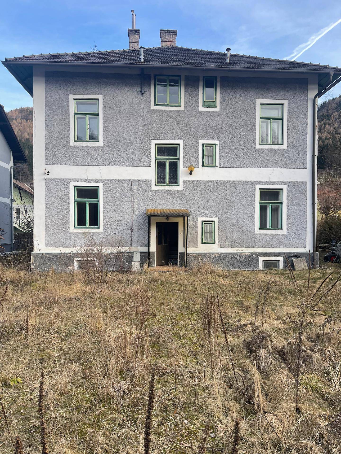 Vorderansicht eines älteren grauen Gebäudes mit vielen grünen Fenstern und einem ungepflegten Vorgarten.