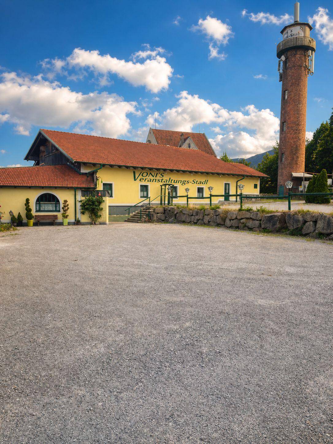 Gesamtansicht des Gebäudekomplexes mit großem Schotterparkplatz und dem hohen Turm im Hintergrund.