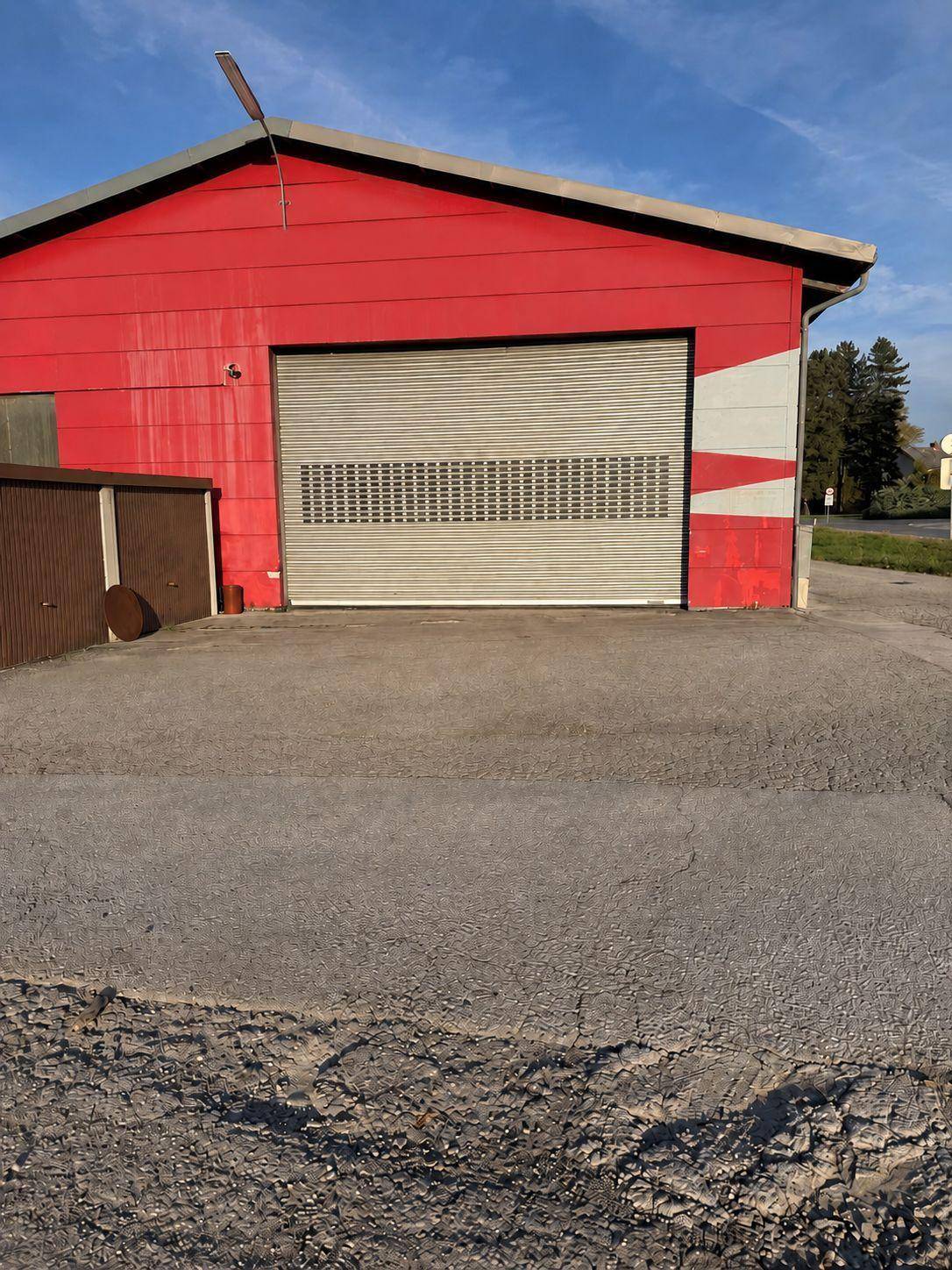 Nahaufnahme einer roten Industriehalle mit einem großen, geschlossenen Rolltor und einem asphaltierten Bereich davor.
