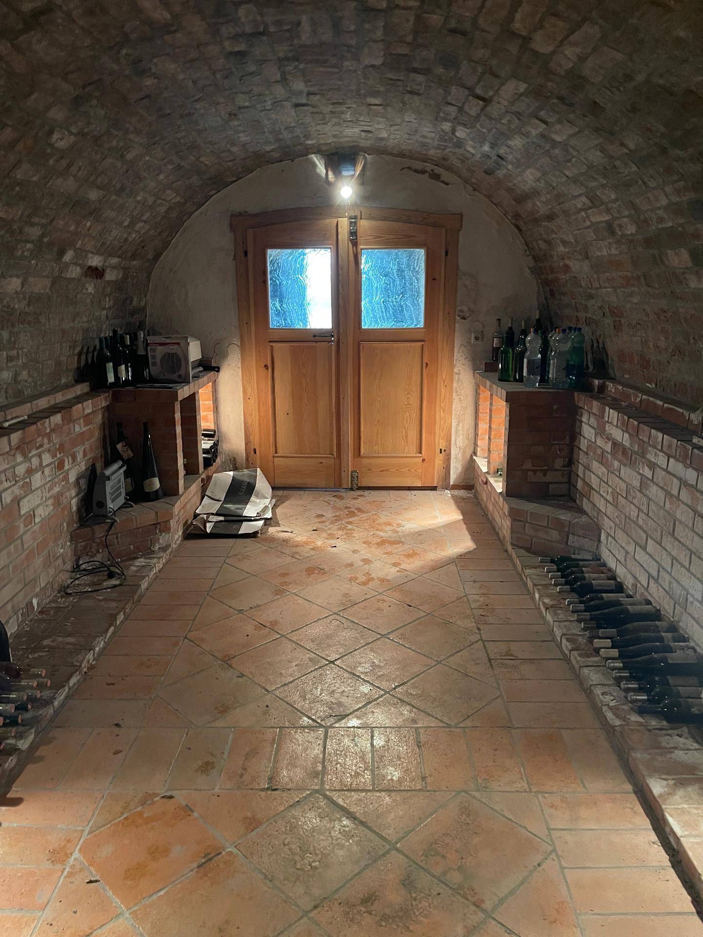 Gewölbter Weinkeller mit Ziegelwänden und Terrakottaboden, gefüllt mit Weinflaschen und einer Holztür am Ende.