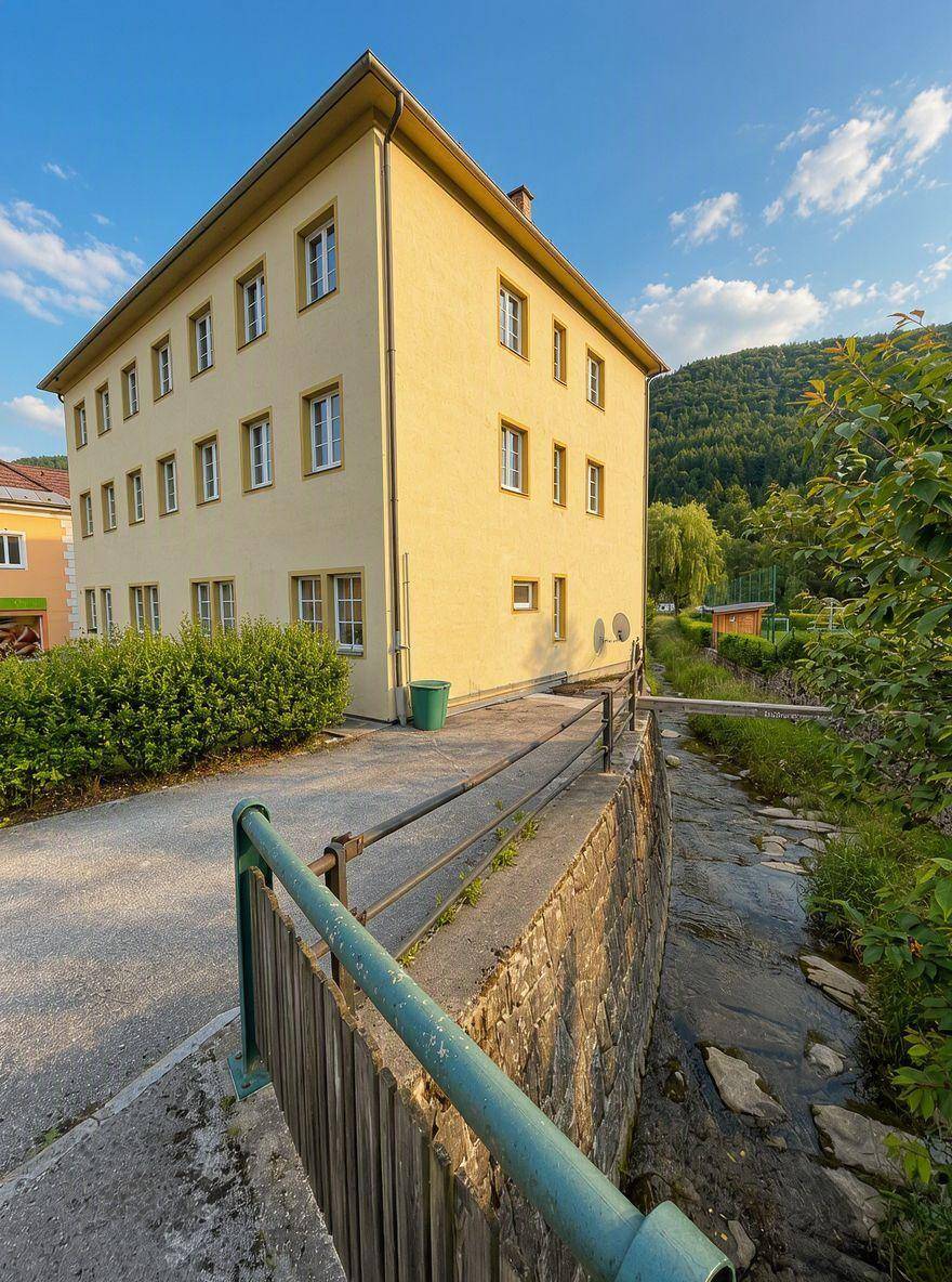 Gelbes Gebäude am Bachlauf mit Blick auf die bewaldeten Berge im Hintergrund.