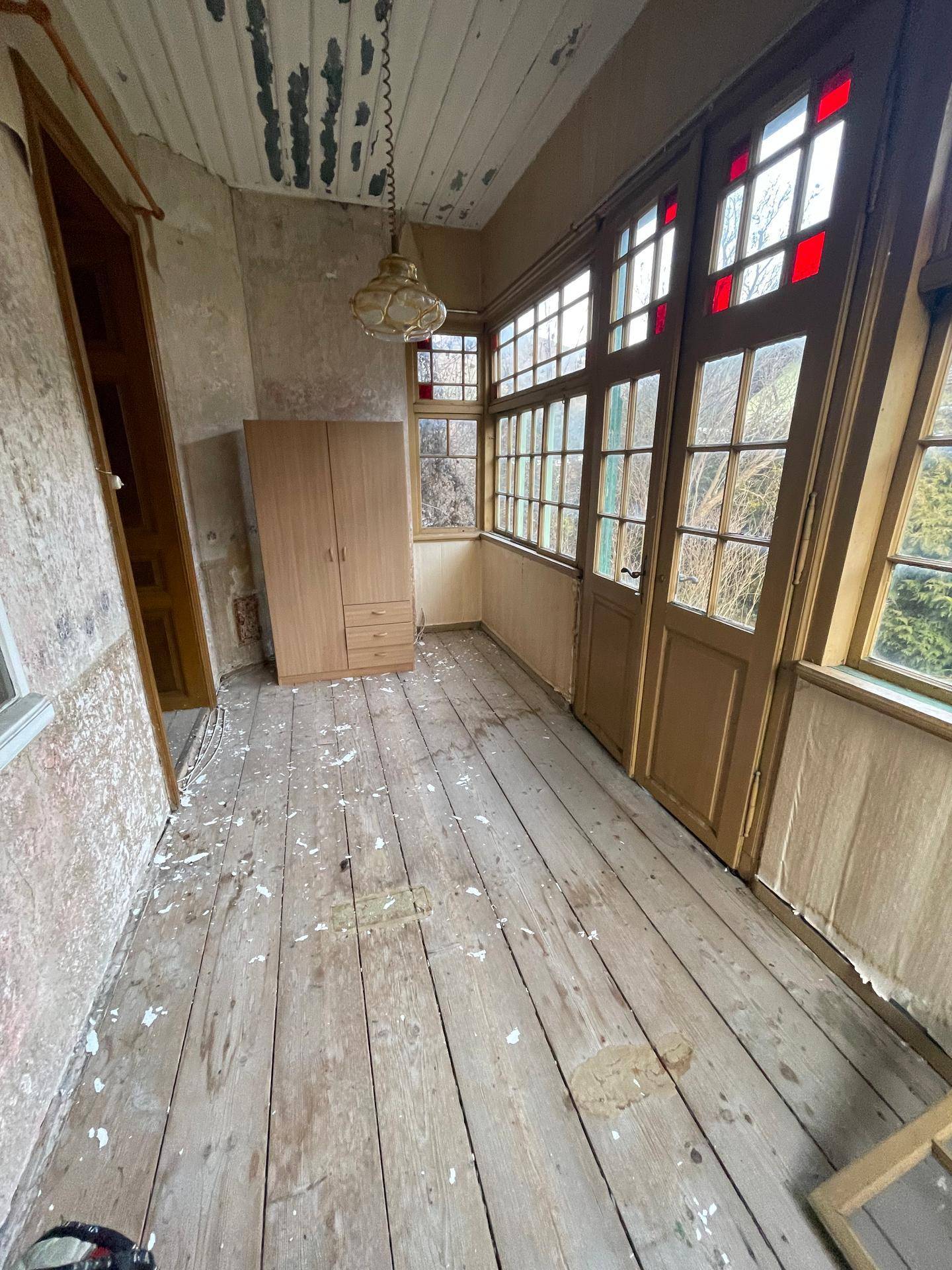 Heller Wintergarten mit Holzplankenboden, vielen Fenstern und einem Kleiderschrank, renovierungsbedürftig.