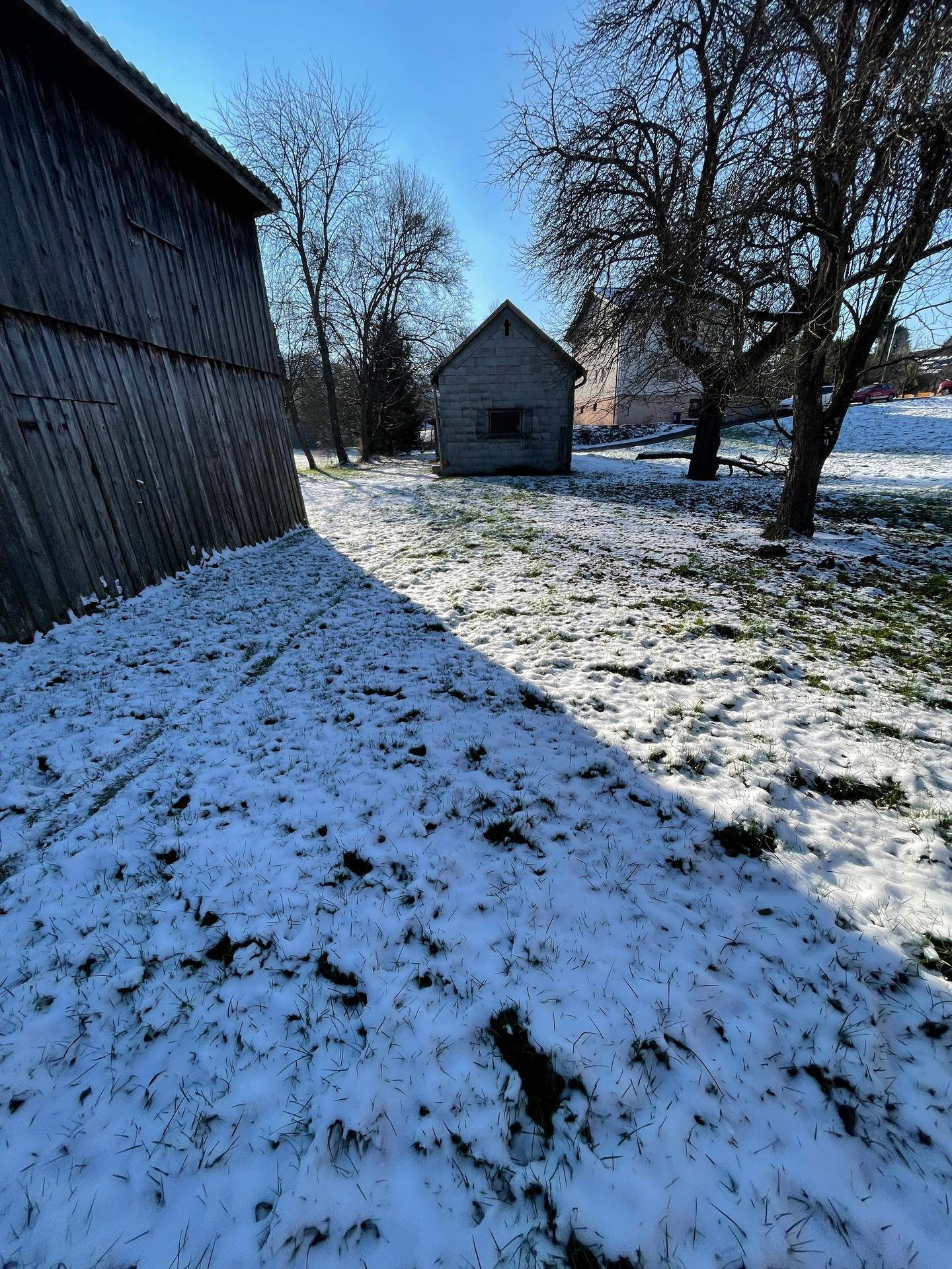 Winterlicher Garten zwischen zwei Holzgebäuden mit einem kleinen Schuppen und kahlen Bäumen im Hintergrund.