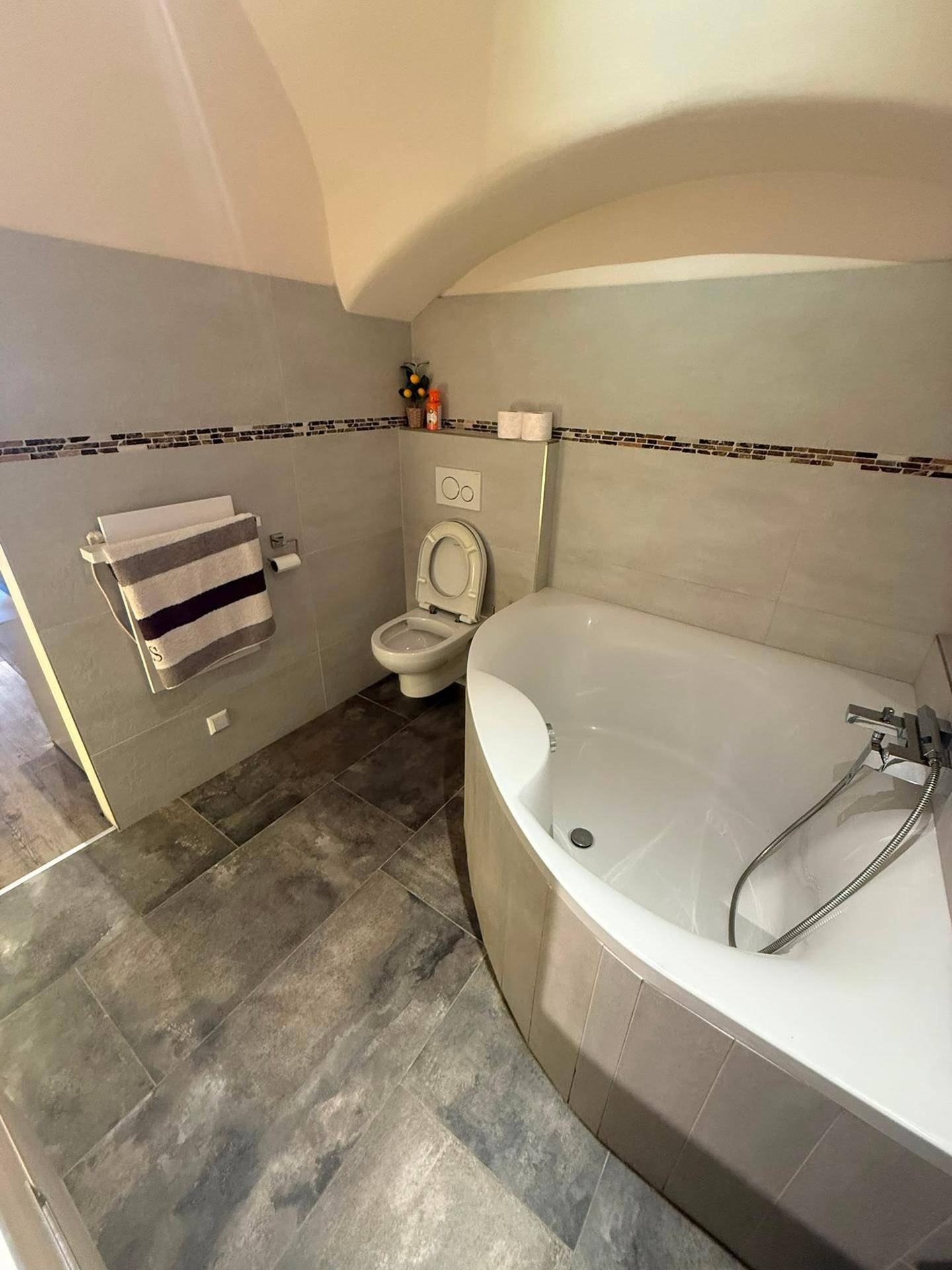 Helles Badezimmer mit Eckbadewanne, wandhängender Toilette und dunklen Bodenfliesen unter gewölbter Decke.