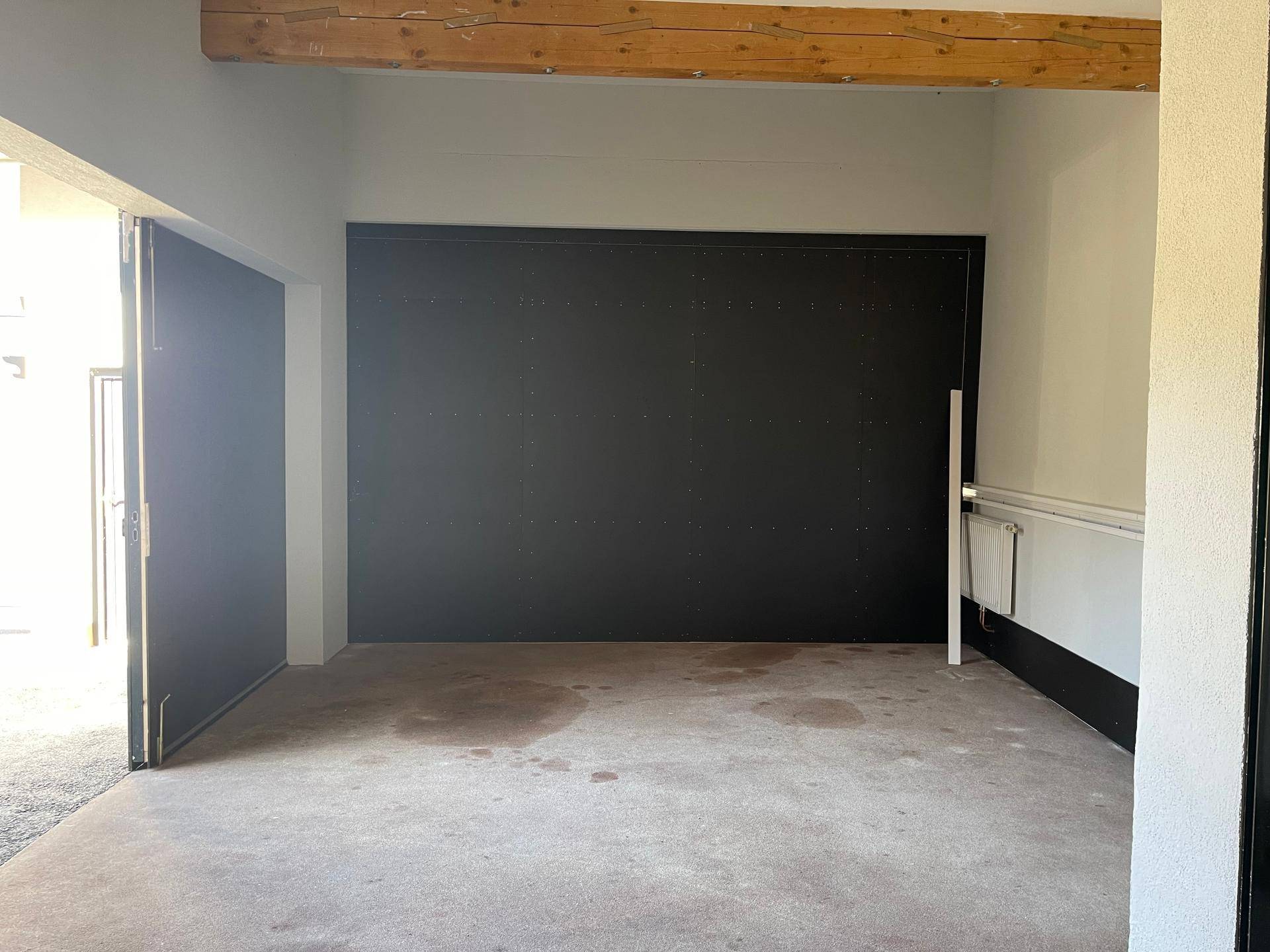 Leerer Garagenraum mit dunkler Rückwand, Betonboden und sichtbaren Holzbalken an der Decke.